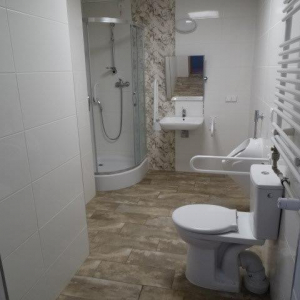 widok łazienki z urządzeniami sanitarnymi (prysznic w kabinie, zlew z lustrem regulowanym, pisuar, sedes toaletowy) i poręczami wspomagającymi